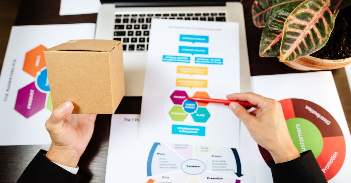 Businessperson holding a cardboard box while reviewing marketing strategy documents at a desk.