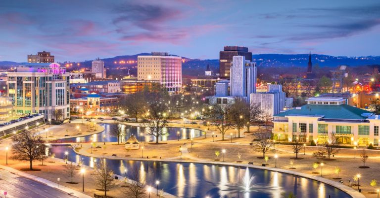 An illuminated city park at dusk with a winding pond reflecting bright park lights and modern downtown buildings under a twilight sky.