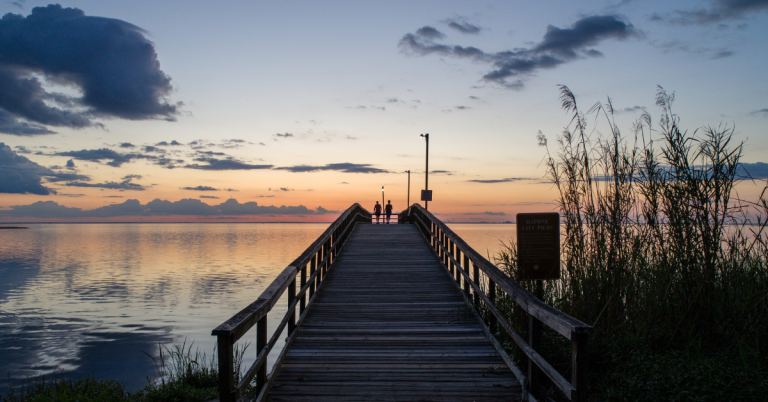 A long wooden fishing pier boardwalk leading out into the calm water of Mobile Bay during a quiet dusk sunset with soft orange and blue clouds in Daphne Alabama.