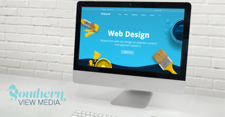 A desktop computer monitor displaying a vibrant blue website design concept template with paintbrushes on a clean white desk against a white brick wall background.